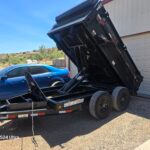 Black IronBull dump trailer with hydraulic lift, parked next to a blue car and garage.